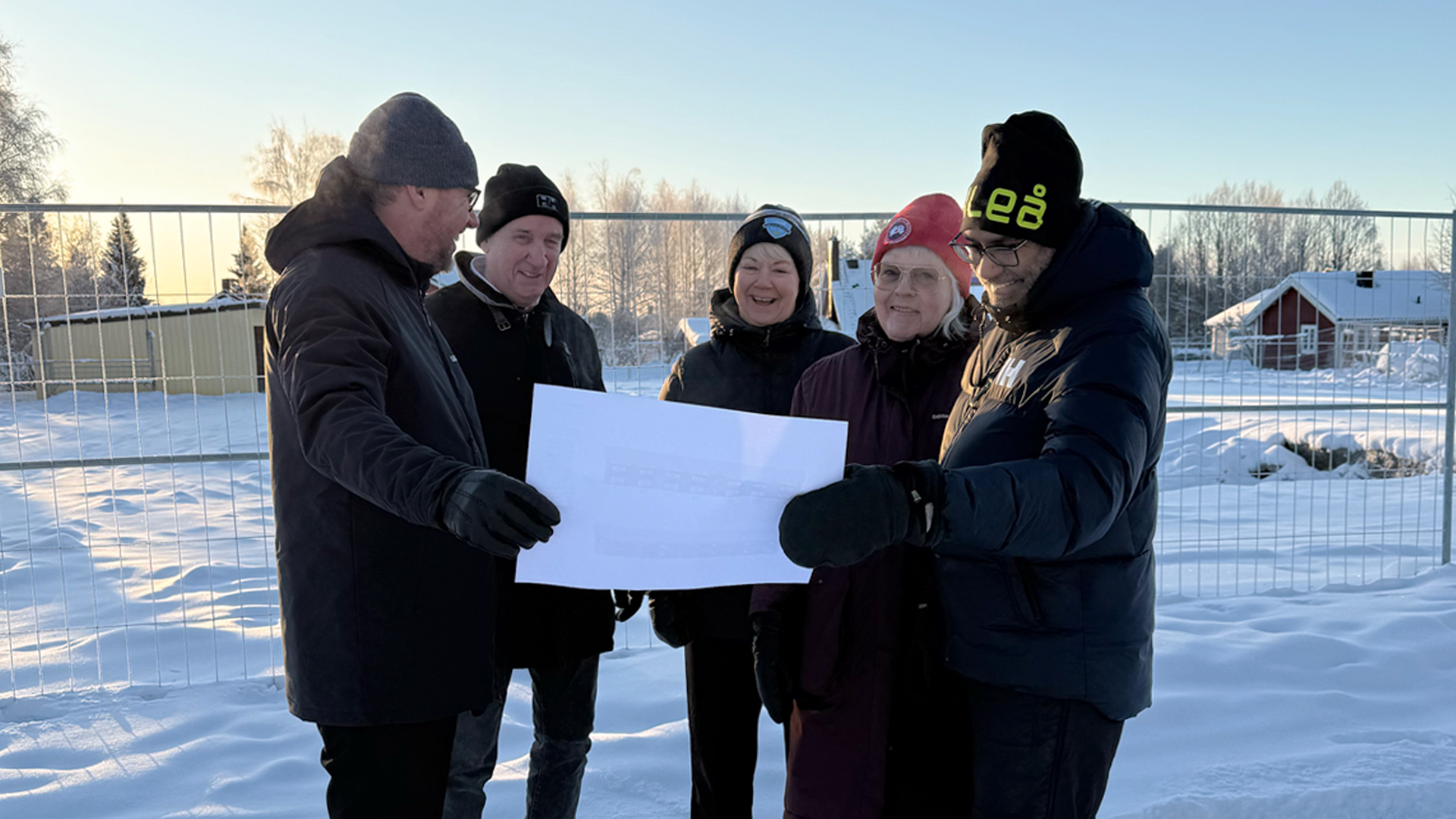 En grupp människor i vinterkläder tittar på ett dokument. De står framför ett byggstängsel som inhä