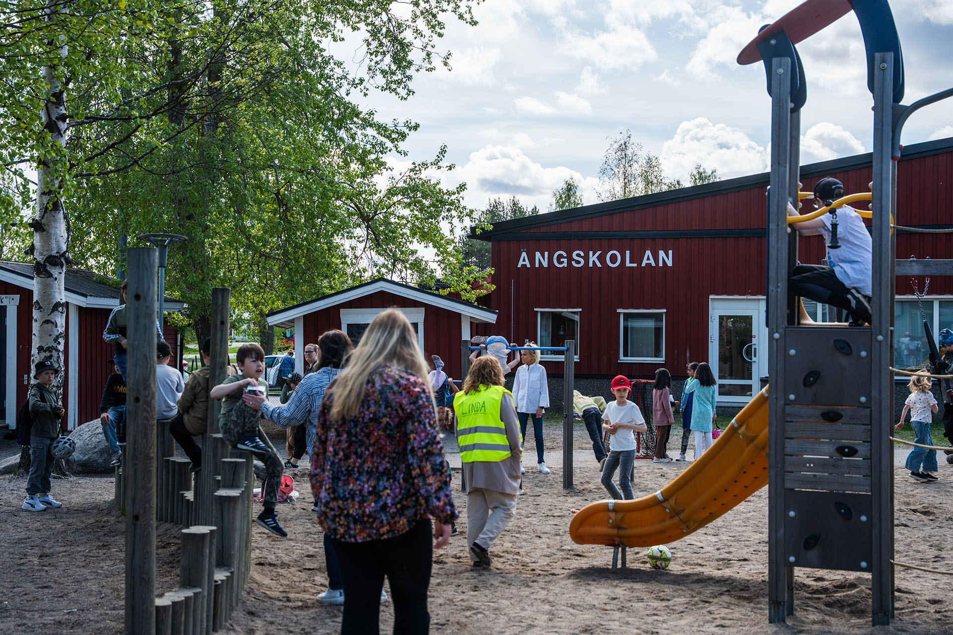Vuxna och barn leker på lekplatsen utanför Ängskolan. I bakgrunden syns skolans fasad.